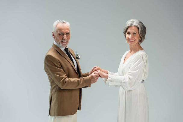happy mature groom and bride smiling while holding hands isolated on grey  - Foto, imagen