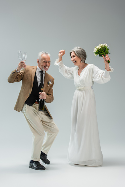 full length of cheerful middle aged man holding bottle of champagne and glasses while dancing with bride in white dress on grey - Fotó, kép