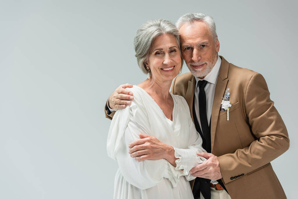 cheerful middle aged man in suit with boutonniere hugging bride in wedding dress isolated on grey  - Fotó, kép