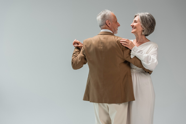 happy middle aged groom and bride in wedding dress holding hands while dancing isolated on grey  - Foto, immagini