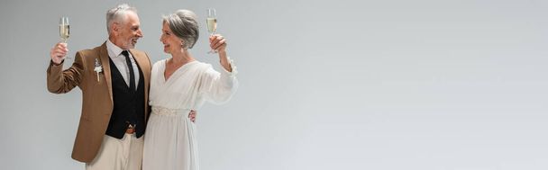 middle aged groom and happy bride in white dress holding glasses of champagne isolated on grey, banner - 写真・画像