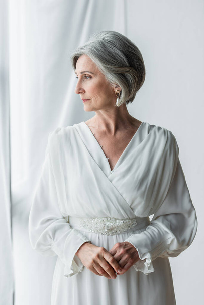 elegant and middle aged bride in white dress standing near curtains and window  - Fotografie, Obrázek