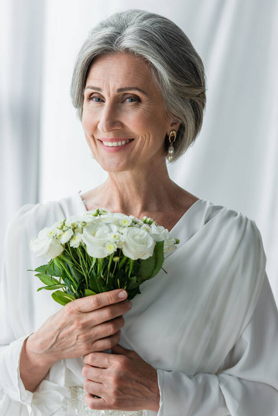 happy middle aged bride in white dress holding wedding bouquet near curtains  - Zdjęcie, obraz