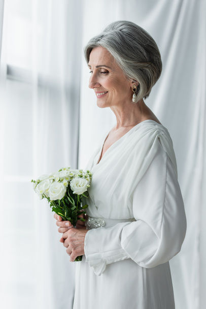 pleased middle aged woman in white dress holding wedding bouquet and standing near curtains - Fotografie, Obrázek