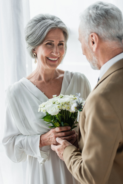 bearded middle aged groom giving wedding bouquet to cheerful bride in white dress - Foto, Bild