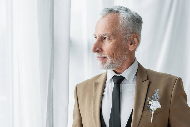 bearded middle aged man in suit with boutonniere looking away near white curtains  - Fotoğraf, Görsel