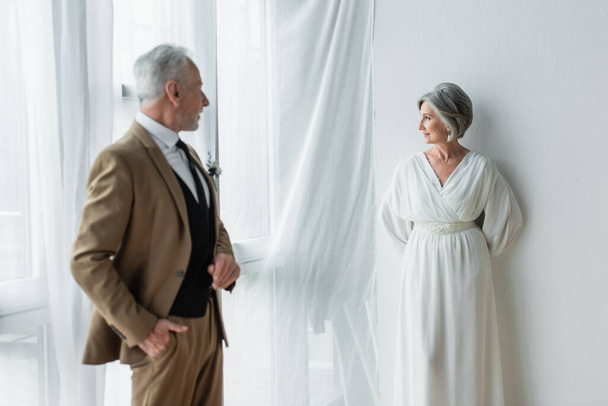 bearded middle aged man in suit posing while looking at cheerful bride in white dress near white curtains  - Foto, immagini