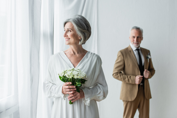 cheerful mature bride in white dress holding wedding bouquet near blurred groom standing near white curtains - Foto, afbeelding