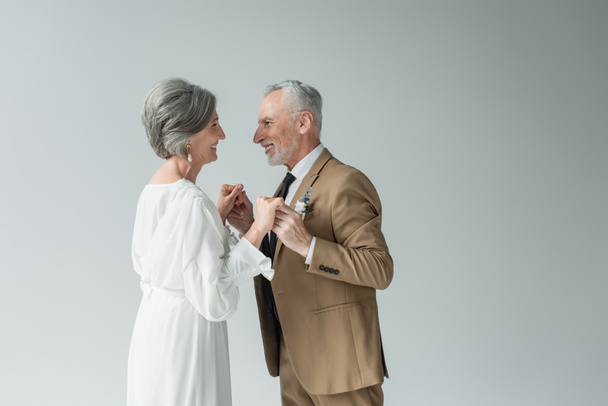 cheerful middle aged man in suit and bride in white wedding dress holding hands isolated on grey - Foto, Imagen