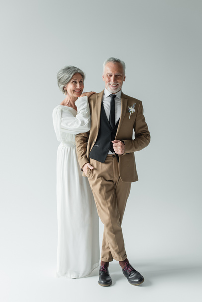 full length of mature bride in white wedding dress and man in suit standing on grey - Foto, Imagem