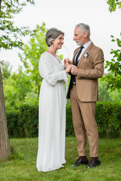full length of happy middle aged groom in suit holding hands with mature bride in white dress in park - Photo, Image
