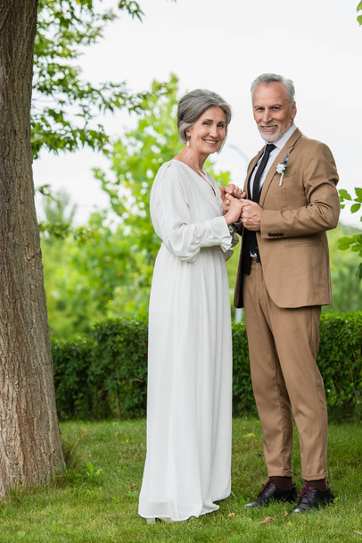 full length of cheerful middle aged groom in suit holding hands with mature bride in white dress in garden  - Fotó, kép