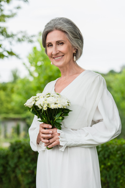 happy middle aged bride in white dress holding wedding bouquet in green garden  - Фото, изображение