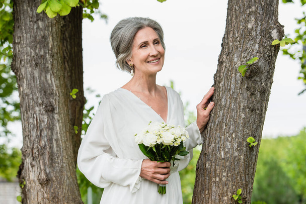 joyful middle aged bride in white dress holding wedding bouquet near tree trunk in park  - Foto, afbeelding