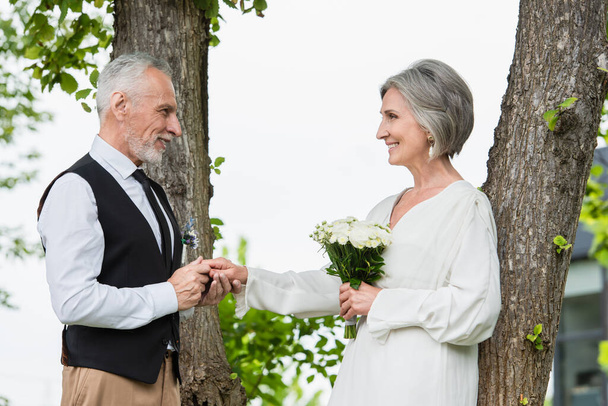 side view of happy mature man in formal wear holding hand of bride with wedding bouquet in garden - Foto, Bild