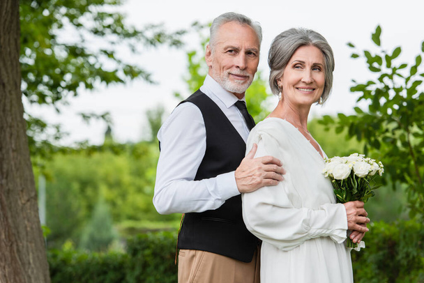 mature man in formal wear hugging smiling bride with wedding bouquet in garden - Photo, image
