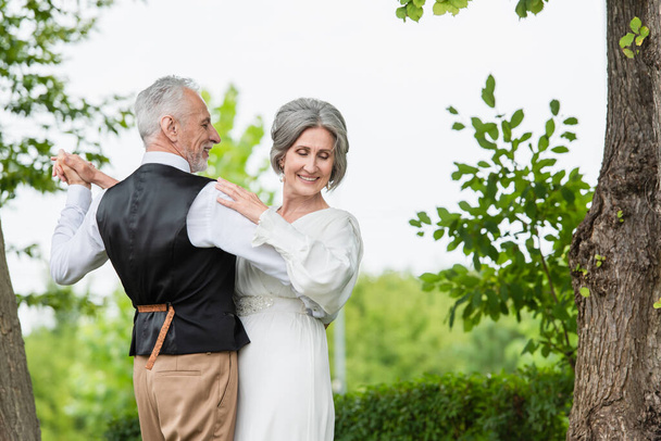 happy middle aged man in formal wear dancing with bride in white wedding dress in green garden  - Foto, afbeelding