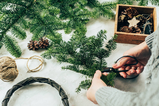 Woman in gray knitted sweater making a christmas fir wreath. Christmas decorating for home interior. Step by step, process. DIY, crafting, hobby concept - Photo, image