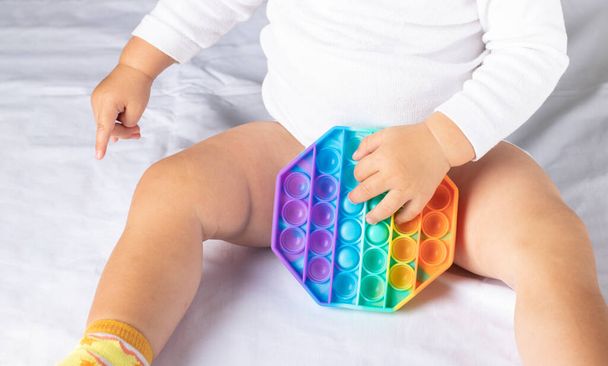 Newborn baby sits on the bed playing trendy flexible silicone colorful toy pop it. Selective focus on anti stress simple dimple toy in childrens hands - Photo, image