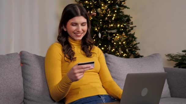 Mujer introduce el código de tarjeta de crédito para pagar regalos de Año Nuevo compras en línea a través de ordenador portátil en el árbol de Navidad de fondo. Compras femeninas de Internet o pagos a través de ordenador sentado en el sofá en casa. Vacaciones de invierno - Metraje, vídeo