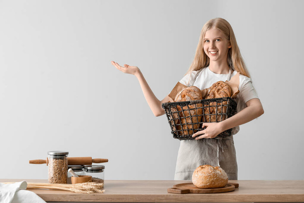 Молодая женщина держит корзину со свежим хлебом и показывает что-то на светлом фоне - Фото, изображение