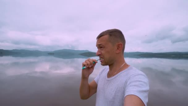 Young man brushes his teeth while standing outdoors on hill with lake and mountains. Oral care in extreme conditions. Picnic or camping in nature in mountains. Soy care. modern tourism. - Footage, Video