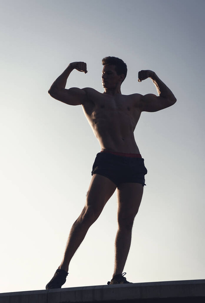 Man showing fit body on blue sky. Bodybuilder posing with muscular hands, biceps and triceps. Athlete standing high on concrete surface. Success and future concept. Fitness and sport. - Fotó, kép