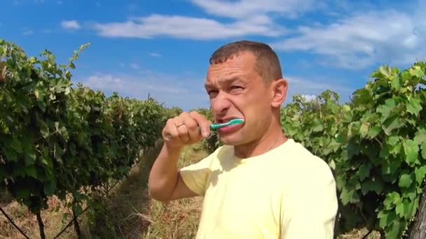 Young man brushes his teeth while standing outdoors against backdrop of vineyards. Oral care in extreme conditions. Picnic or camping in nature. Self-care. modern tourism. - Video