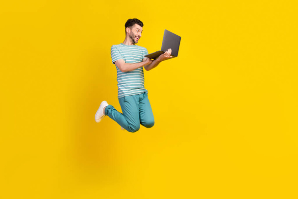 Full length photo of impressed excited man dressed striped t-shirt blue pants jumping look on laptop isolated on yellow color background. - Foto, Imagen