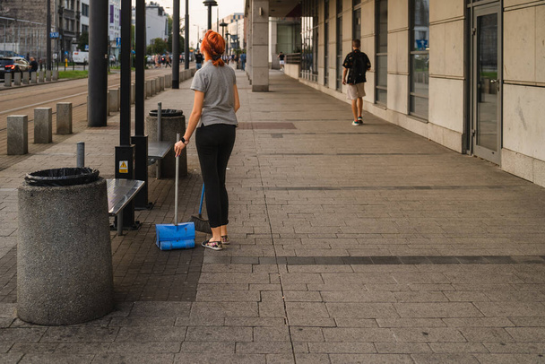 A girl in casual clothes sweeps garbage at a city tram stop in the evening, a girl with a dustpan and a broom stopped and looks dreamily into the distance, street photography, youthful dreams - Photo, Image