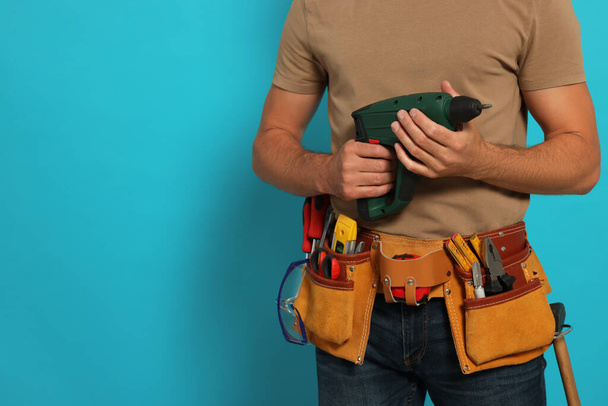 Professional builder with tool belt and power drill on light blue background, closeup. Space for text - Zdjęcie, obraz