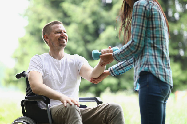 Женщина-физиотерапевт помогает молодому человеку в инвалидном кресле поднять вес рук в парке. Реабилитация инвалидов и физической активности - Фото, изображение