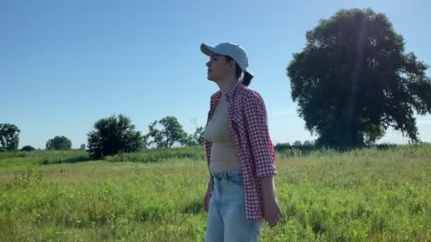 Νεαρή ενήλικη γυναίκα περπατά στο λιβάδι την αυγή. Όμορφη γυναίκα που φοράει καπέλο, καρό πουκάμισο, τζιν. Εξοχή τρόπο ζωής. Γυναίκα αγρότισσα πηγαίνει σε αγροτική γη. Κινούμενη βολή - Πλάνα, βίντεο
