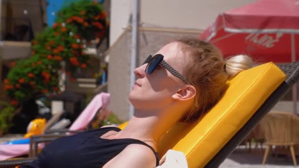 Woman in sun protective glasses and black swimsuit lies on chaise longue near pool, relaxing, sunbathing. Summer holidays, vacations, concept of travel. Caucasian blonde tans. - Filmmaterial, Video