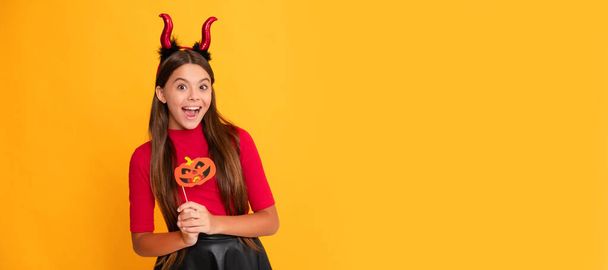 glad teen girl wearing imp horns holding pumpkin party accessory. Halloween kid girl portrait, horizontal poster. Banner header with copy space - Fotó, kép