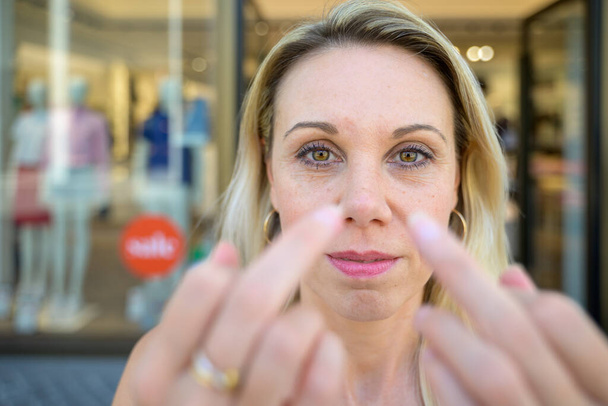 Middle aged blond woman posing in front of urban shops raising her middle fingers to her face in a close up portrait - Foto, afbeelding