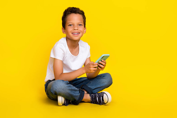 Full length photo of little boy sit hold telephone wear t-shirt jeans boots isolated on yellow color background. - Foto, Bild