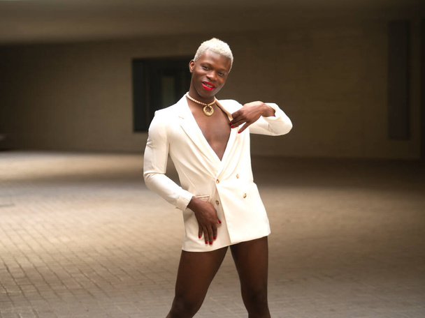 Glad androgynous African American man with short blond hair and red lips looking at camera with smile and touching elegant white jacket while standing in spacious room - Foto, immagini