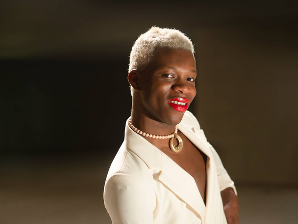 Cheerful African American androgynous man in stylish white jacket with red lips and short blond hair holding hand on waist and looking at camera with smile in dark room - Zdjęcie, obraz