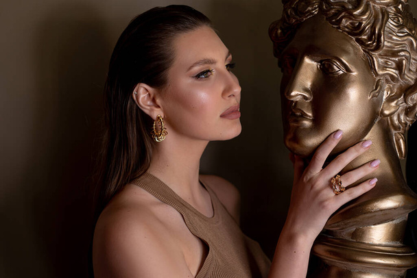 Portrait of young flawless thoughtful woman with long dark hair, bright golden make-up, pink lips touching head face of bronze bust David by Michelangelo on grey background. Beauty, cosmetics, art. - Foto, Imagen