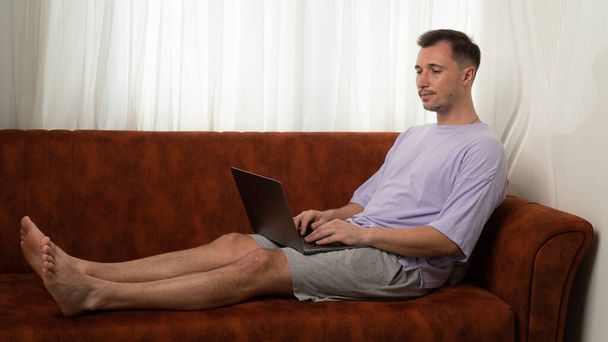 Un hombre yace en el sofá y escribe en una computadora portátil, trabajando en la computadora en casa. Foto de alta calidad - Foto, imagen
