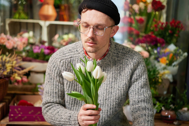 Portrait of attractive bearded male in gray sweater, eyeglasses and beanie hat holding beautiful bouquet of spring white tulips. Mothers Day, Valentines or International Womens Day concept - Foto, Imagem