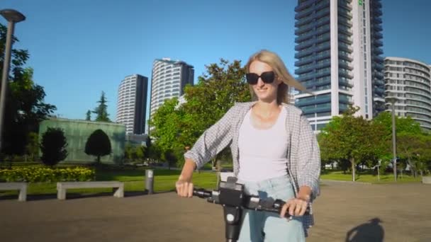 Young beautiful girl rides electric scooter in summer along street, against background of city and park, she is happy and very pleased with ecological and fast mode of transport. Summer, sunny day, - Footage, Video