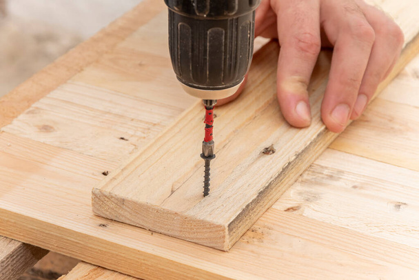 Κοντινές λεπτομέρειες ενός Καυκάσιου αρσενικού ξυλουργού χρησιμοποιώντας ένα ηλεκτρικό κατσαβίδι στο χέρι του και επισκευάζοντας ένα νέο ξύλινο τραπέζι, η έννοια της οικιακής βελτίωσης - Φωτογραφία, εικόνα