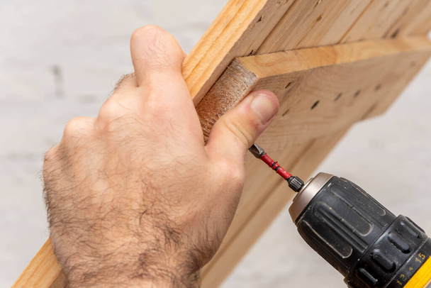 A close-up of an old electric screwdriver in his hand, a screw is being screwed into a new wooden table. Concept: home improvement, furniture repair with your own hands. - Photo, image