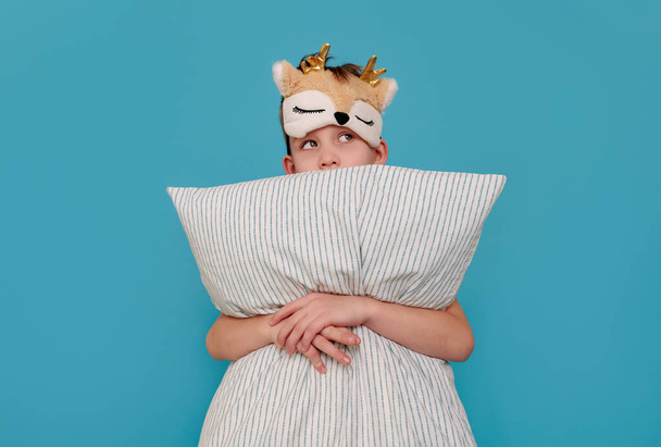 the cunning boy does not want to sleep, hugs the pillow and looks away. Child on a blue background with copy space. The concept of a naughty child, parenting, insomnia, health - Foto, Bild