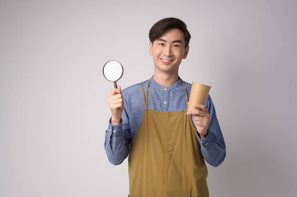 A Portrait of young asian man wearing apron holding magnifying glass and paper cup over white background studio. - Foto, Imagen
