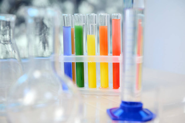 Focus on test tubes with rainbow colored chemicals and reagents standing on tripod. Blurred foreground with graduated cylinder, flasks and measuring glass labware. Chemistry, science concept - Zdjęcie, obraz
