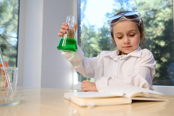 Adorable little girl has fun doing chemistry experiments, shakes a flat-bottomed flask with green solution, reads the steps of doing a science experiment. Development of children from an early age - Zdjęcie, obraz