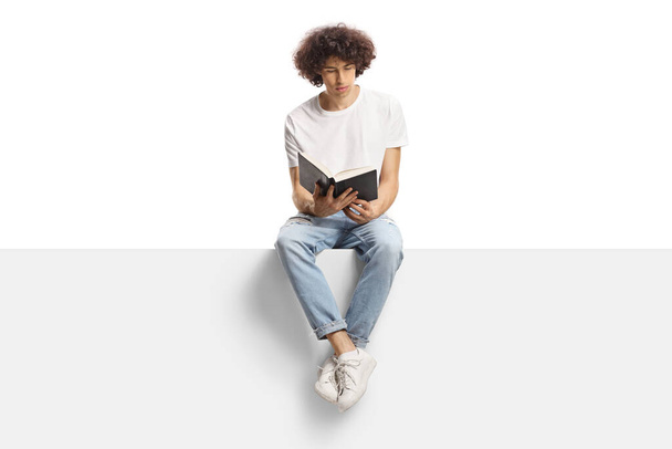 Young man with curly hair sitting on a panel and reading a book isolated on white background - 写真・画像